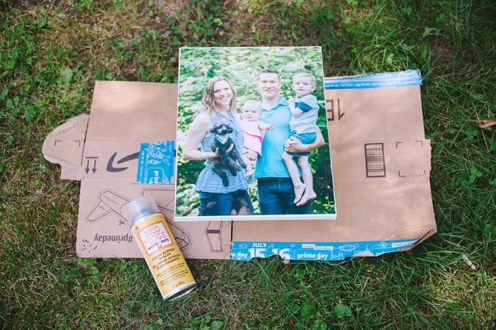 A handmade photo canvas laying outside on the grass, on top of a flattened cardboard box, with a can of clear acrylic sealer next to it.