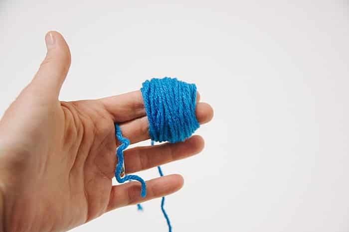 A hand with yarn wrapped around two fingers, in the process of making a pom pom.