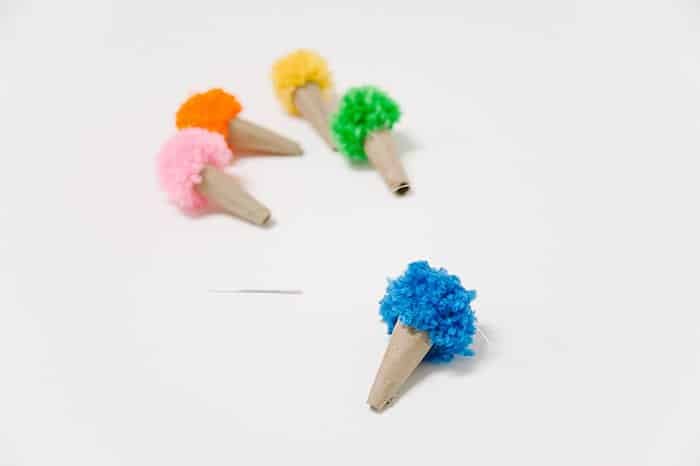 Colorful ice cream cones with cardboard cones and yarn pom pom tops laying next to fishing line and an embroidery needle.