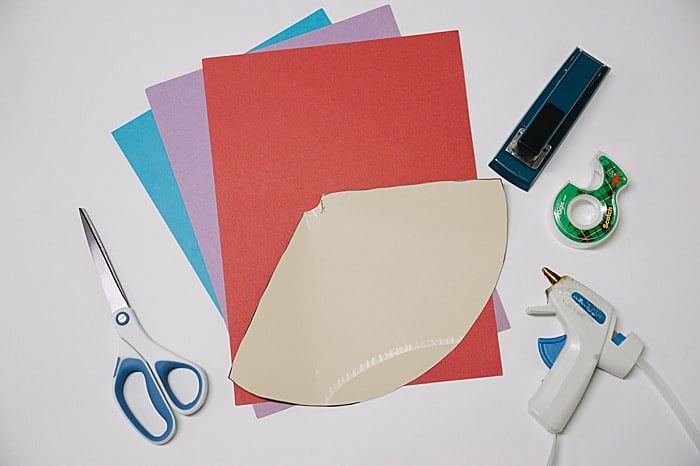 Supplies needed to make a cone out of paper including construction paper, a template for a cone, scissors, tape, stapler and hot glue gun.