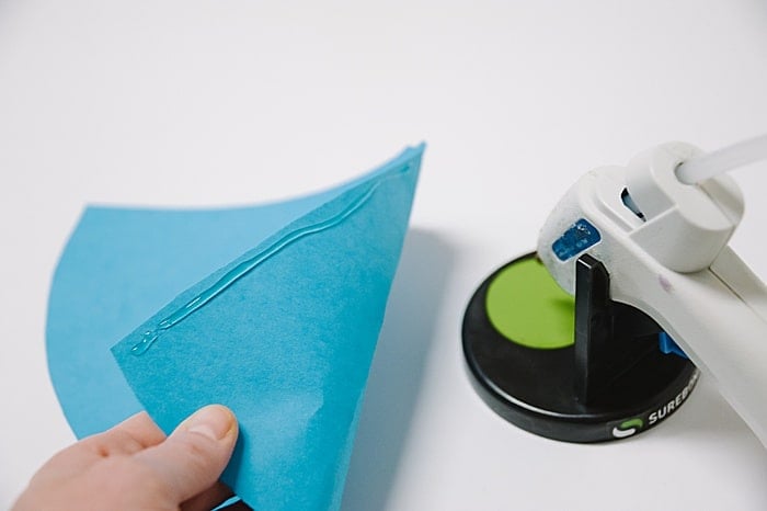 A blue paper cone with hot glue along the edge, and a hot glue gun in a stand nearby.