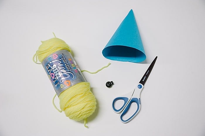 Supplies to make a paper party hat, including a yellow skein of yarn, scissors, a black cord lock stopper, and a handmade blue paper cone.