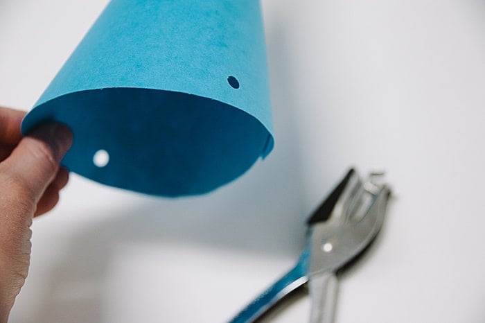 Blue paper party hat with two holes punched in the sides, and a single hole punch laying on the table next to it.