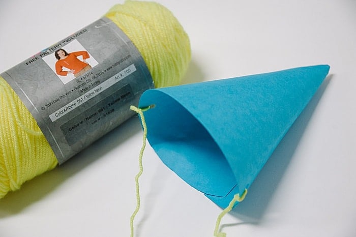 A skein of neon yellow yarn laying next to a DIY blue paper party hat with yellow yarn strings.