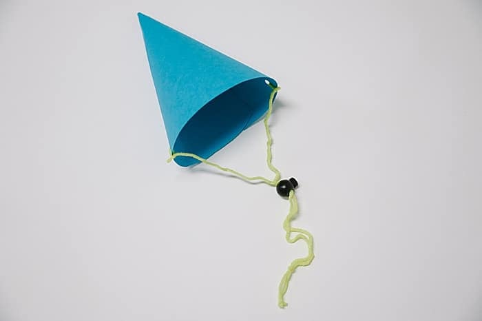 A handmade blue party hat with a yellow string and black stopper laying on a white surface.