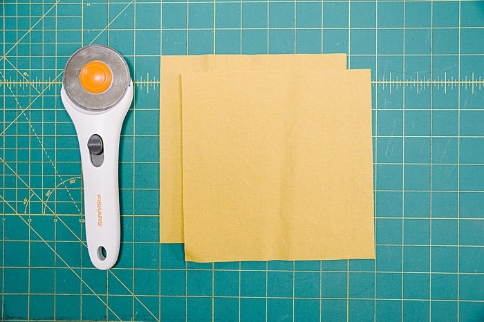 Two yellow 7 inch by 7 inch squares for cotton duck fabric laying on a green rubber cutting mat next to a rotary cutter.