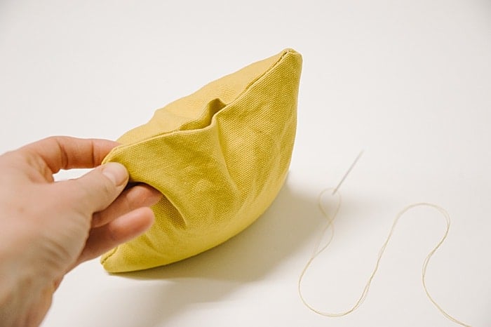 A yellow handmade cornhole bean bag with filling added through an open turn hole, with a needle and thread laying nearby.