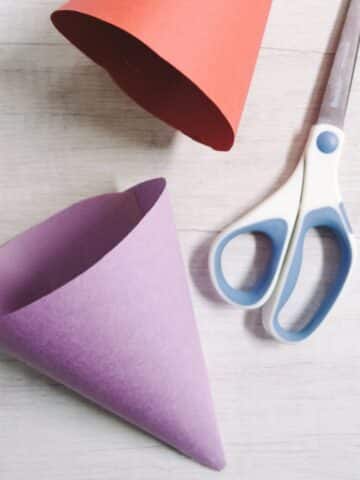 Purple and red paper cones made with construction paper, laying with a scissors on a table top.