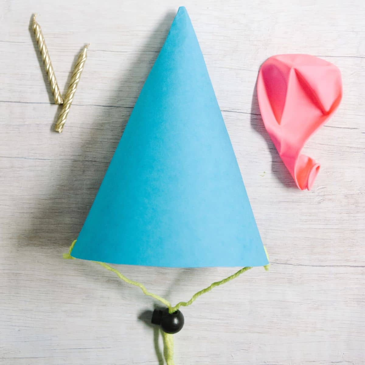 DIY paper party hat made with construction paper, yarn, and a cord lock stopper. Laying on a table top with birthday candles and a pink deflated balloon.