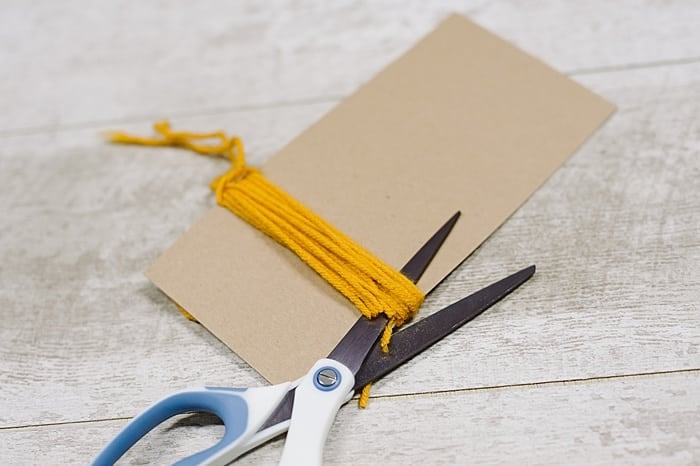 A scissors cutting the bottom of a yarn tassel as it's wrapped around cardboard.