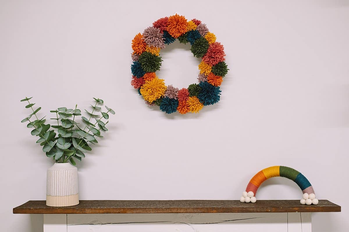 A rainbow pom pom wreath hanging on the wall above a wood fireplace.