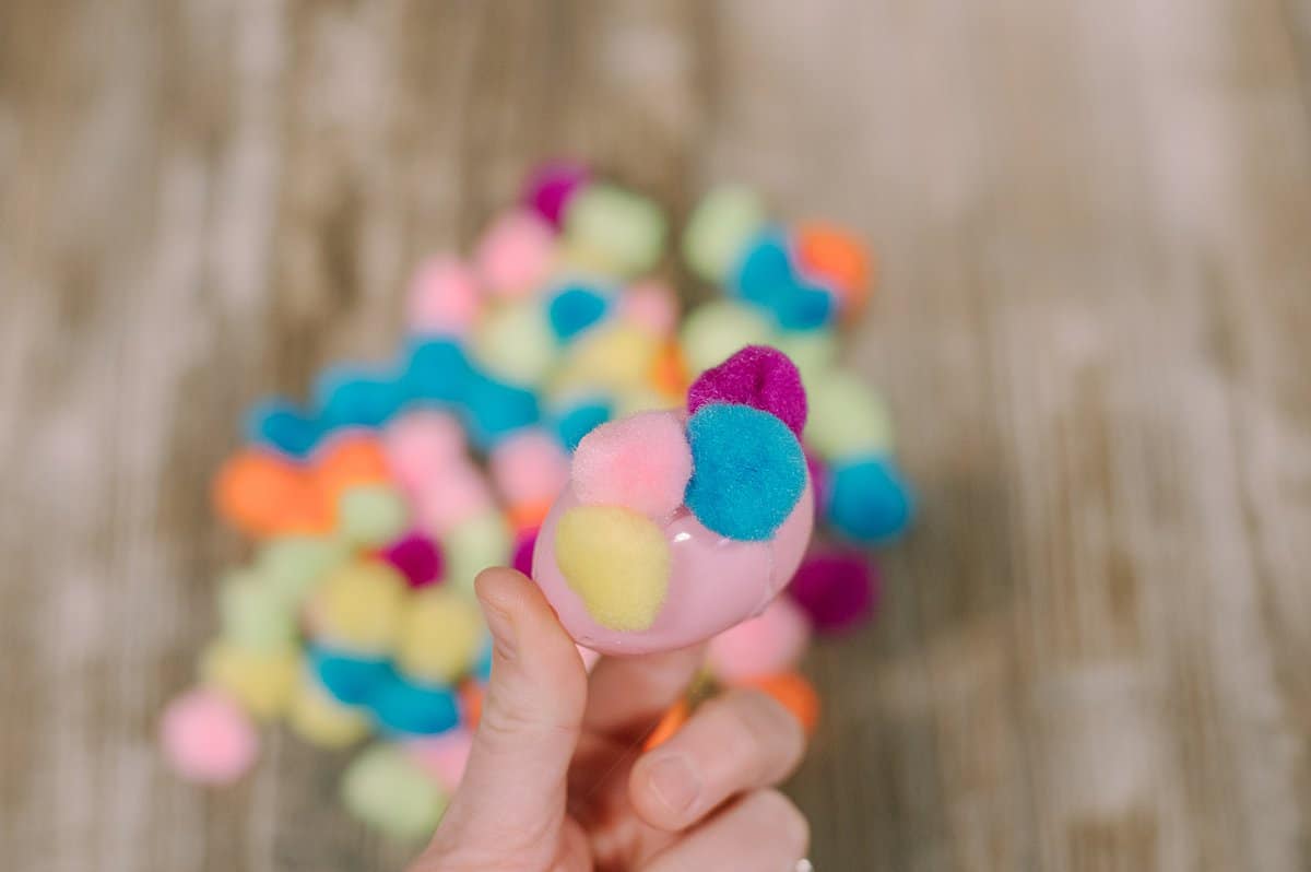 A pink plastic egg with pom poms being hot glued around the outside of the egg.