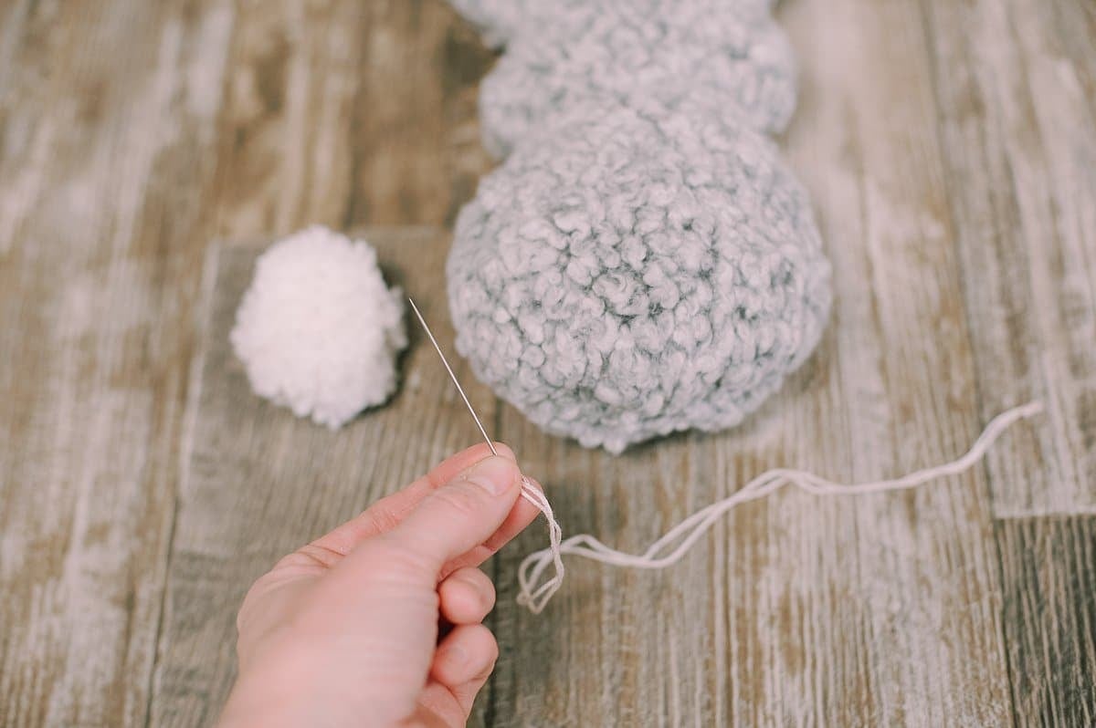 A bunny shaped stuffed animal with a yarn pom pom laying next to it. An embroidery needle, embroidery floss, and a hand holding the needle.