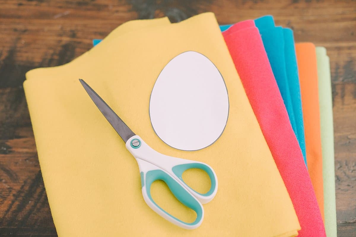 Pieces of felt in bright spring colors, with a paper egg shape and pair of scissors laying on top of the pile.