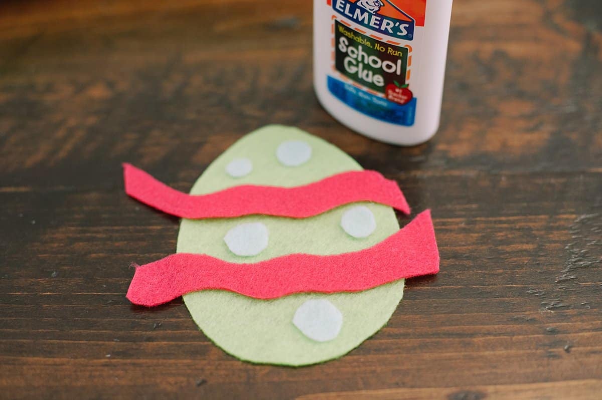 A green felt egg shape with felt stripes and circles being glued onto it with school glue.