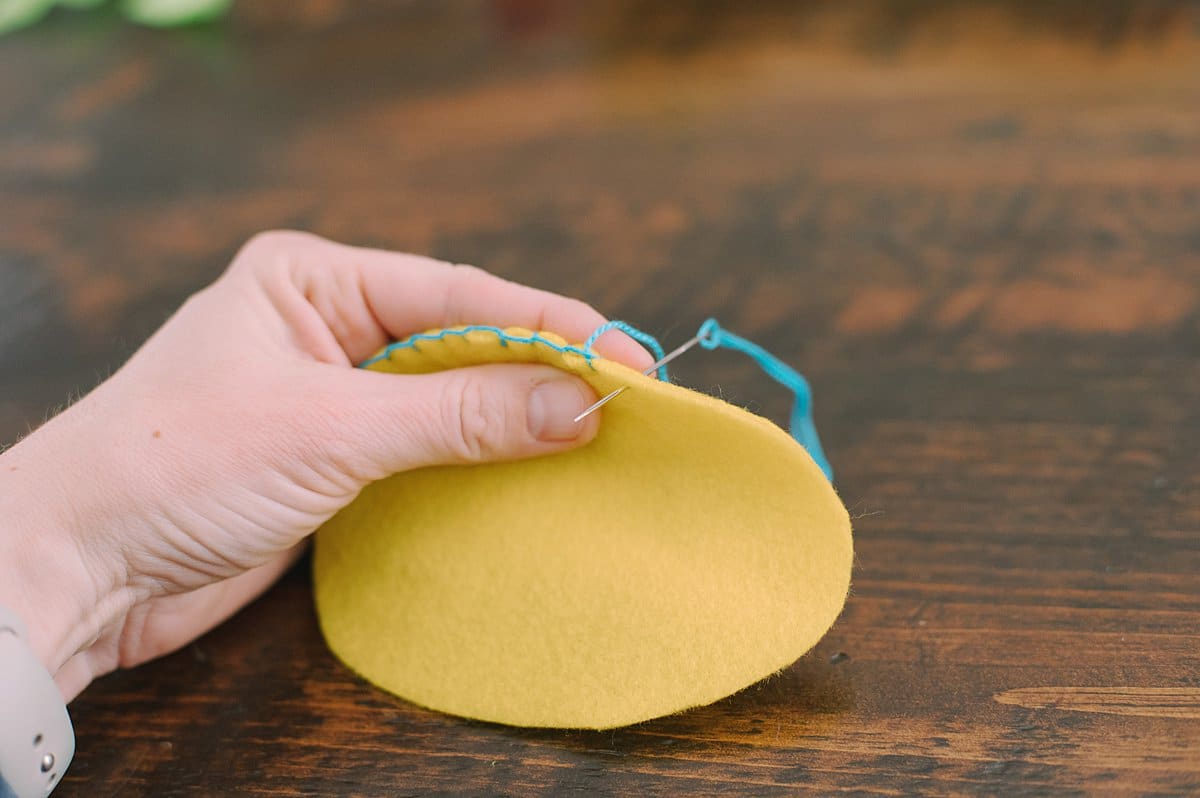 A hand holding egg shaped felt and stitching the outside edge with a needle and thread, doing the blanket stitch.