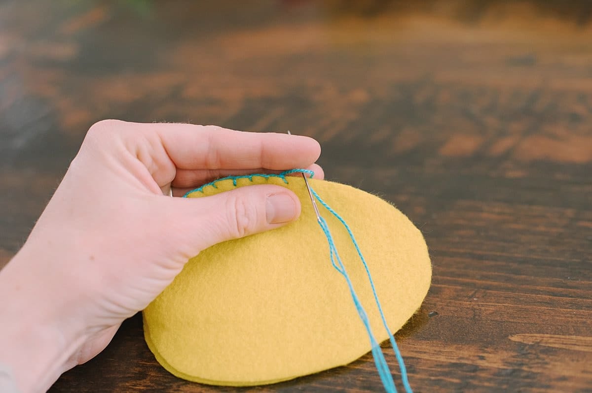 A hand holding egg shaped felt and stitching the outside edge with a needle and thread.