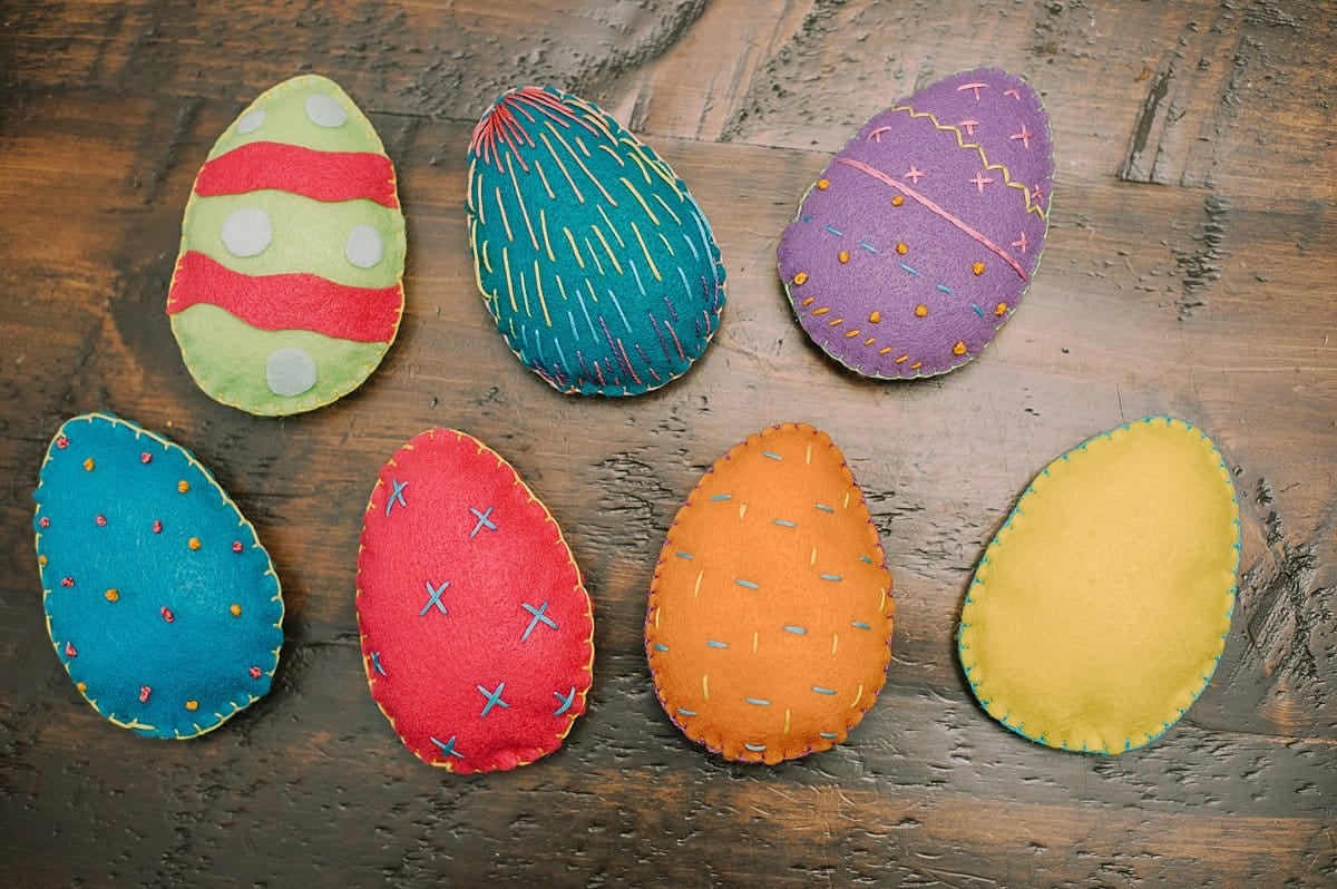 Felt easter eggs in rainbow colors with embroidery stitches on the front, laying on a wood surface.