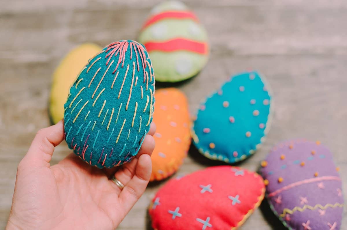 A hand holding a teal felt easter egg with multi-colored straight embroidery stitches on the front.