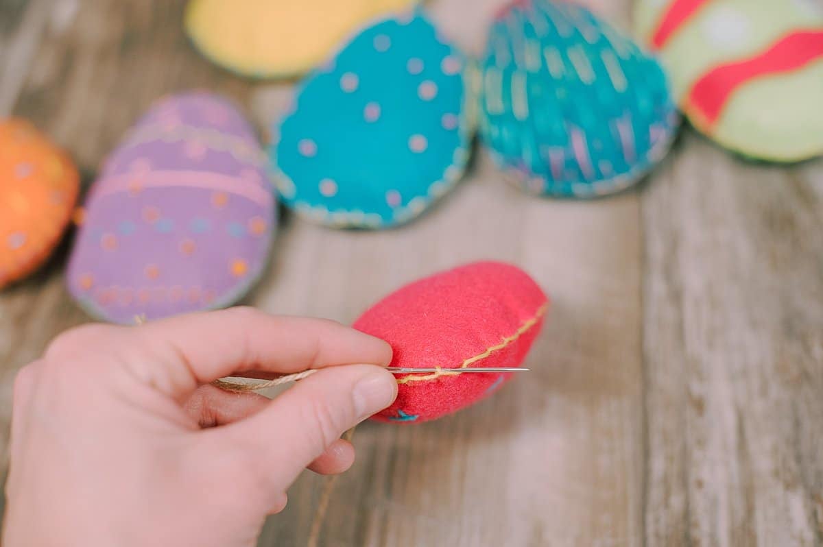 A needle and twine string being threaded through the embroidered edge of a felt Easter egg.