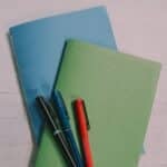 Two DIY paper books with blue and green covers laying on a tabletop with three pens on top of them.