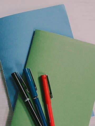 Two DIY paper books with blue and green covers laying on a tabletop with three pens on top of them.