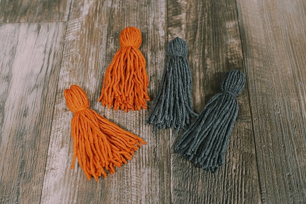 Orange and gray yarn tassels with freshly trimmed, straight bottoms, laying on a wood floor.