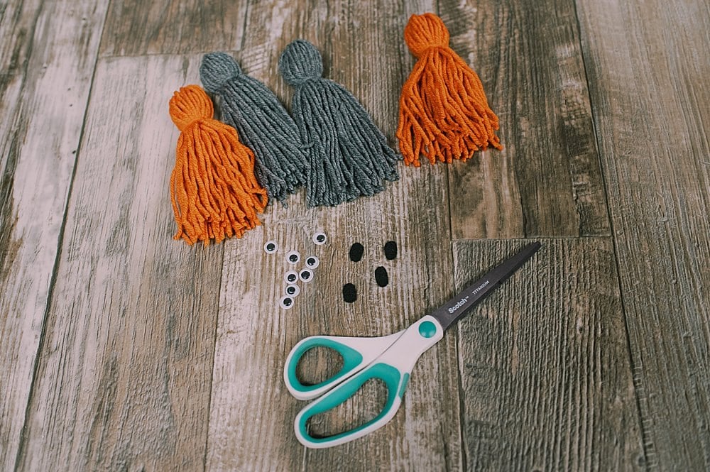 Four yarn tassels laying next to googly eyes, small felt circles, and a scissors.