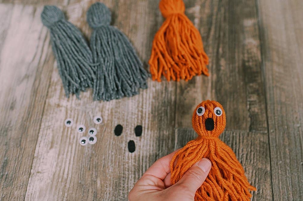 An orange yarn tassel with googly eyes and a black felt oval mouth hot glued onto the head of the yarn tassel to make a ghost.