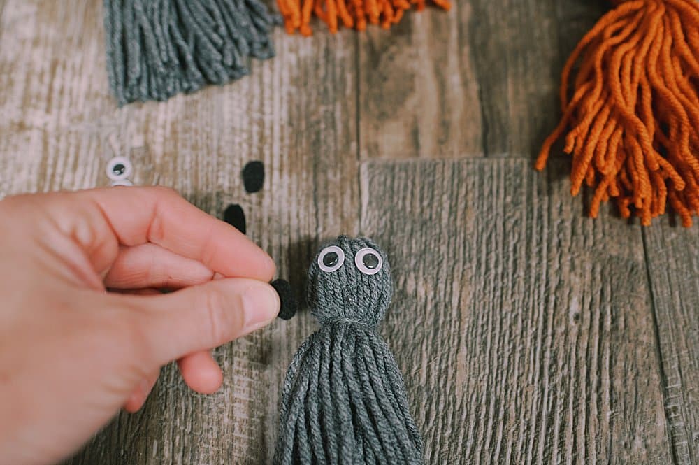 A yarn tassel with two googly eyes glued onto it, and a hand with a small black circle of felt nearby.