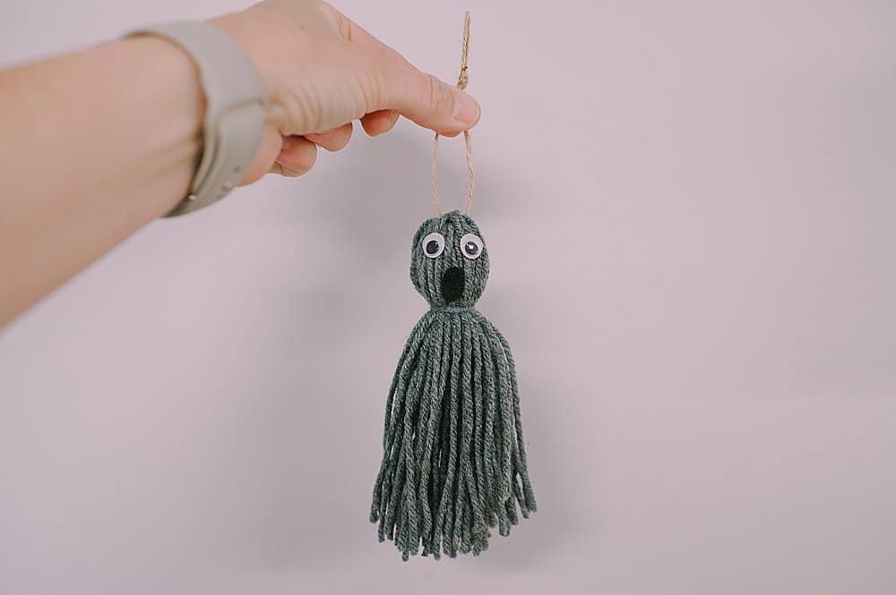 A gray yarn tassel ghost with a twine hanging string, help up by a hand.