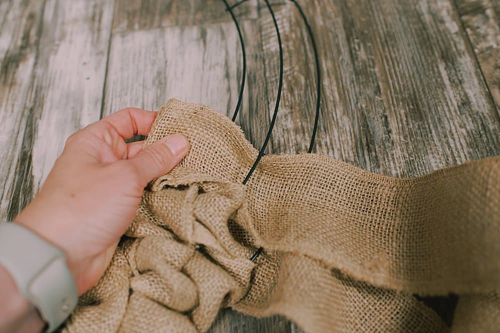 A hand weaving burlap ribbon in and out of a wire wreath form.