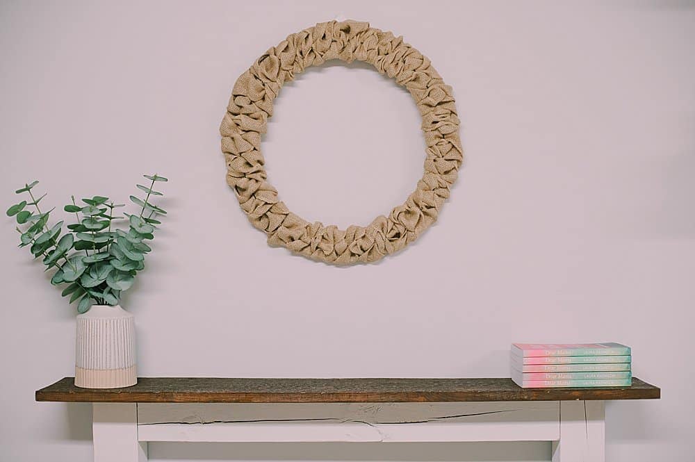 A rustic wreath made with burlap ribbon hanging above a wood mantel with a plant and a stack of books on the mantel.
