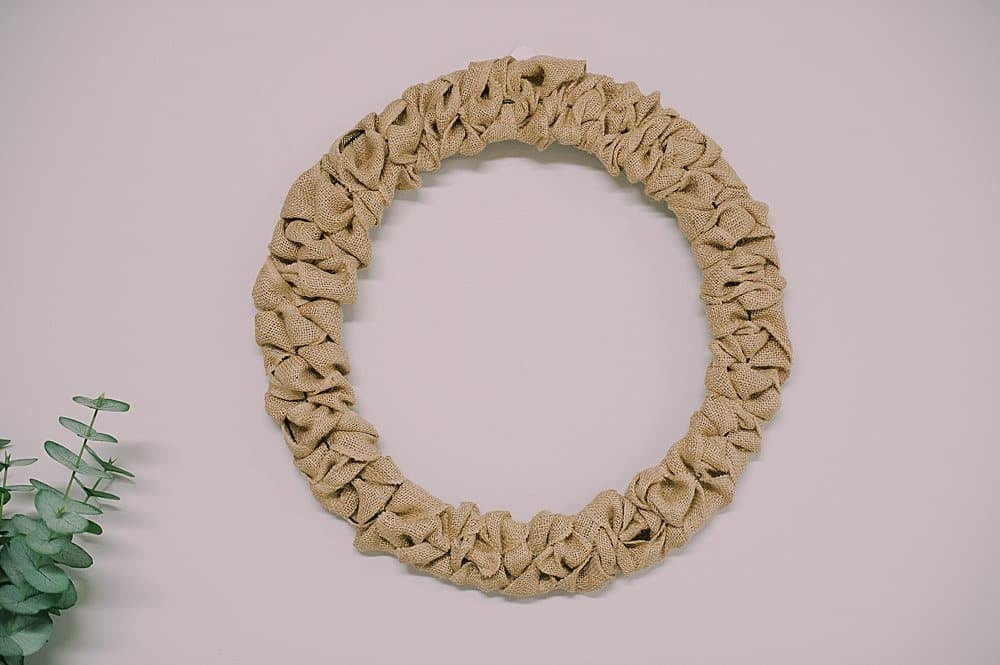 A wreath made with burlap ribbon hanging up on a wall next to a green plant.