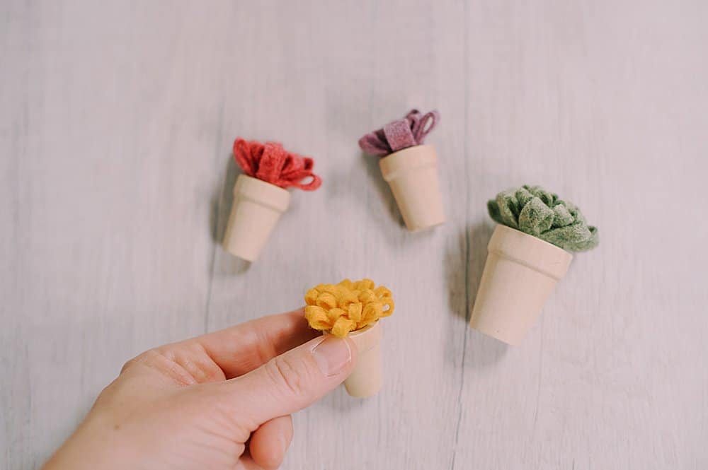 Tiny felt mum flowers inside wooden pots, laying on a white wood surface.