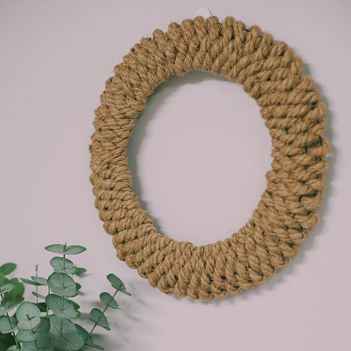 Photo showing a wreath made out of jute rope hanging on a wall above a shelf with a plant on it.