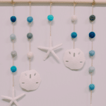 How to make a DIY ocean wall hanging using felt balls, faux sand dollars and starfish, twine, and a wooden dowel.