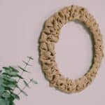 A rustic wreath made with burlap ribbon and a wire wreath form having on a wall next to a fireplace mantel and plant.
