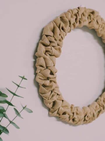 A rustic wreath made with burlap ribbon and a wire wreath form having on a wall next to a fireplace mantel and plant.
