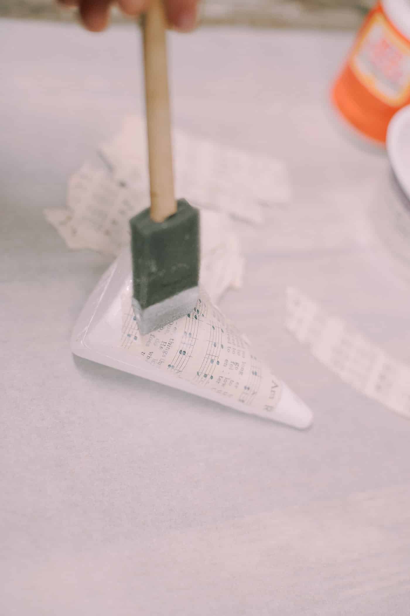 A styrofoam cone with Mod Podge on it, and a brush coating a piece of book page onto the surface of the cone.